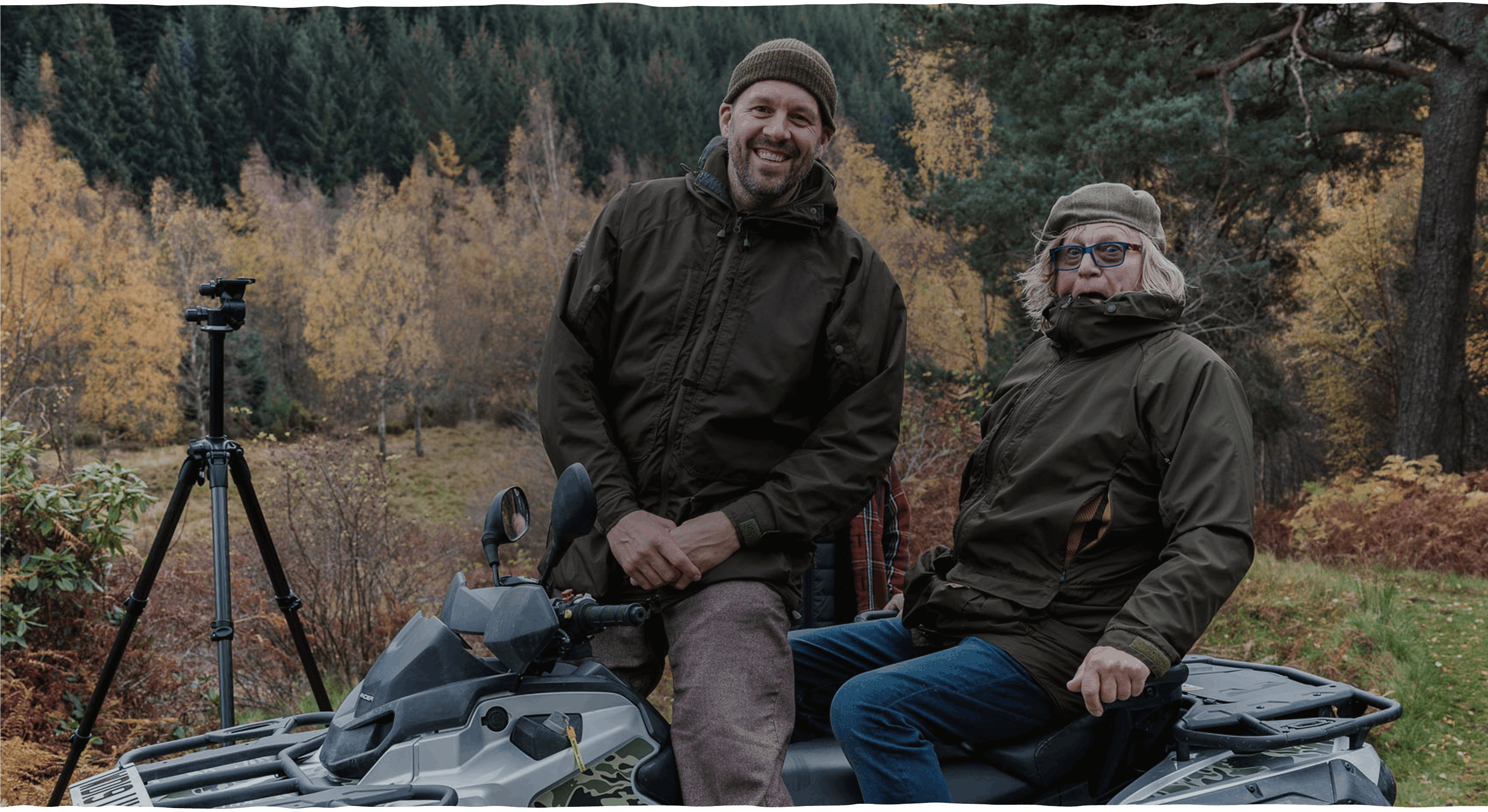 Two people wearing brown jackets sit on an all-terrain vehicle in a forested area with autumn foliage in the background
