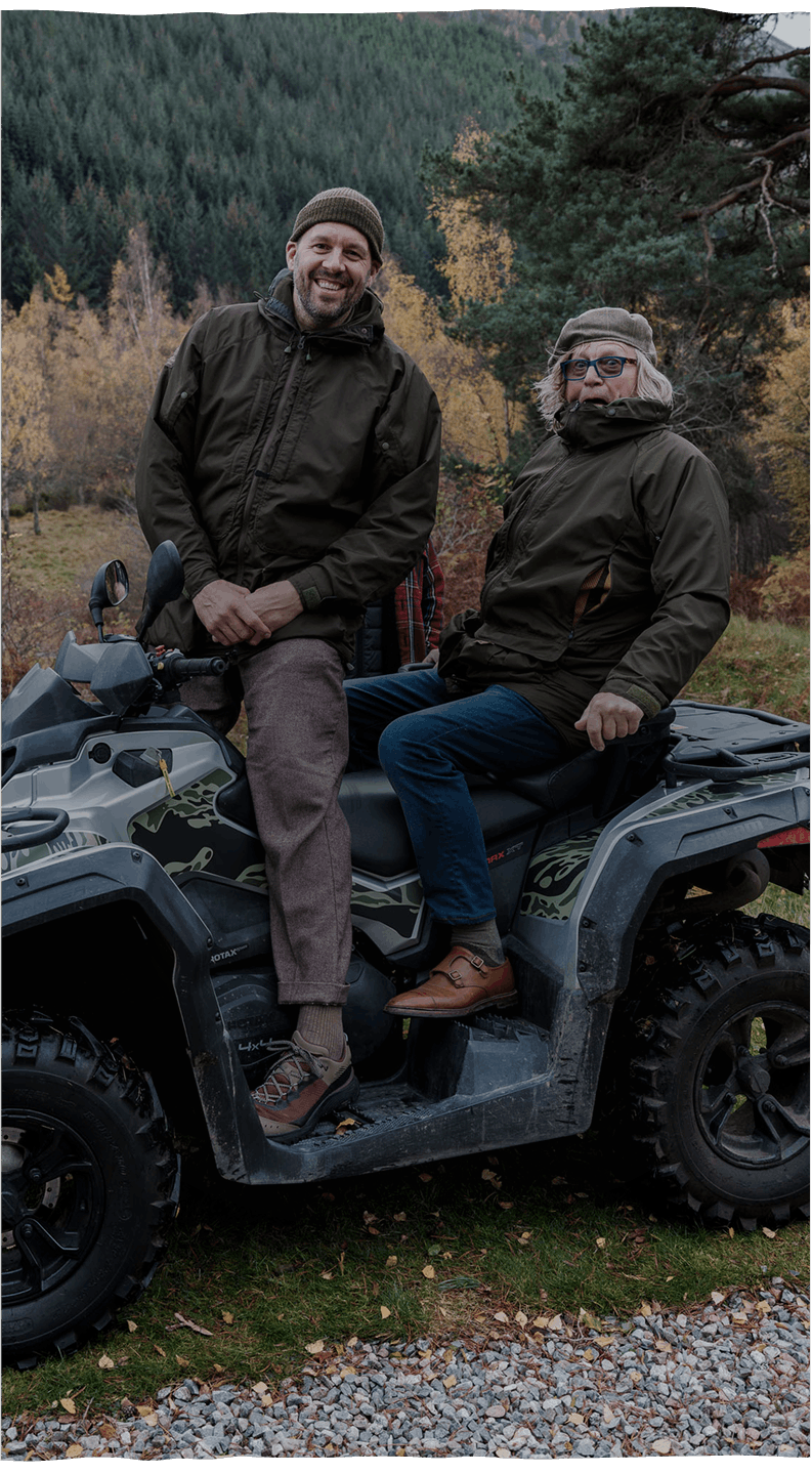 Two men in outdoor jackets sitting on a camo ATV, surrounded by autumn foliage and gravel path in a forest setting