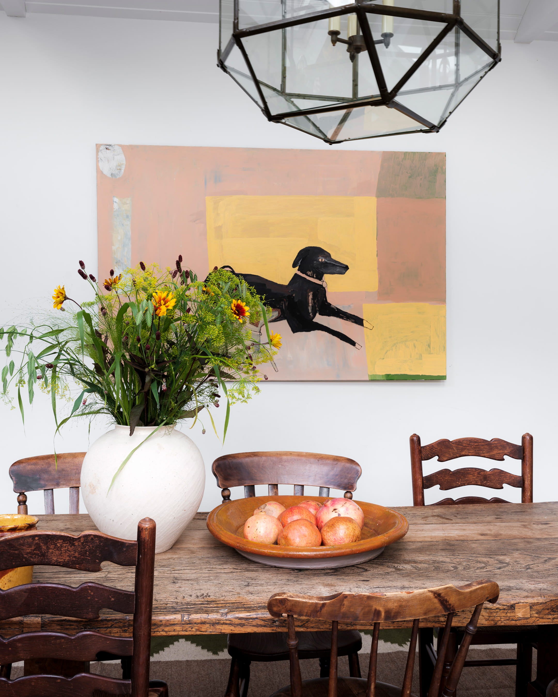 Rustic dining area featuring a reclaimed wood table, vintage wooden chairs, a large white vase with wildflowers, and abstract artwork