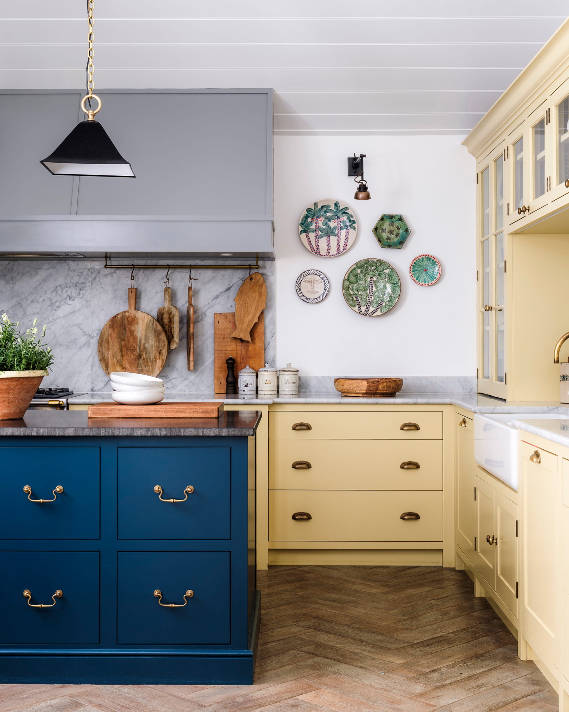 Bright kitchen featuring a deep blue island, pale yellow cabinetry, herringbone oak flooring, and decorative ceramic plates on the wall