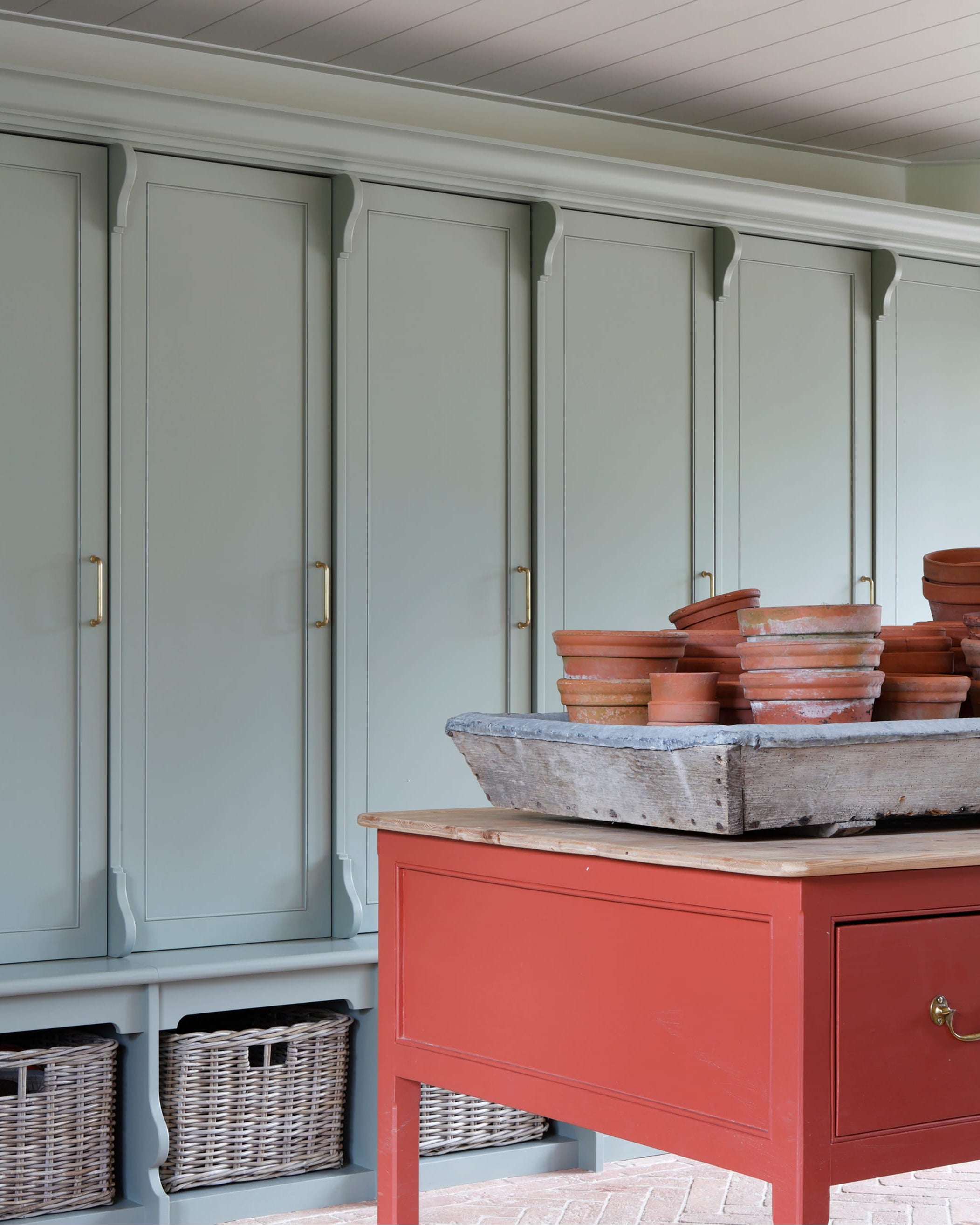 Muted sage green cabinetry with copper handles, a rustic red table, and terracotta pots atop a weathered metal tray
