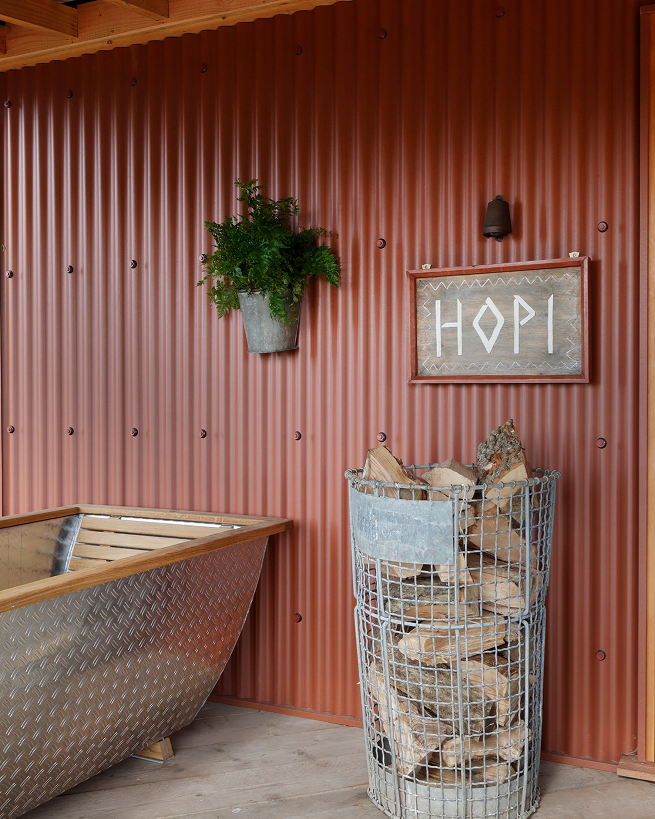 Rustic interior featuring a corrugated rust-red wall, a wooden bathtub, and a wire log basket filled with firewood