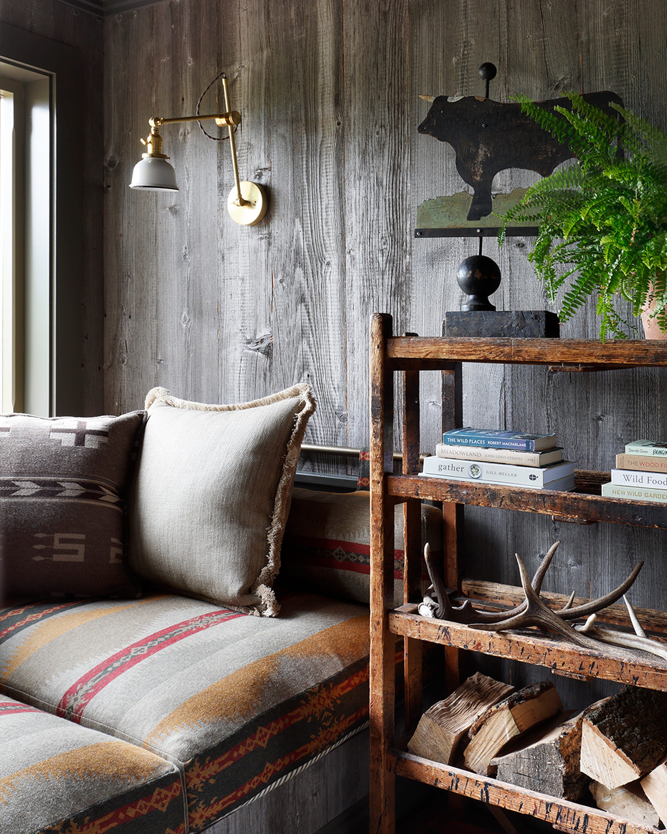 Cozy nook with weathered wood walls, a patterned textile seat, and a rustic wooden shelf displaying books and antlers