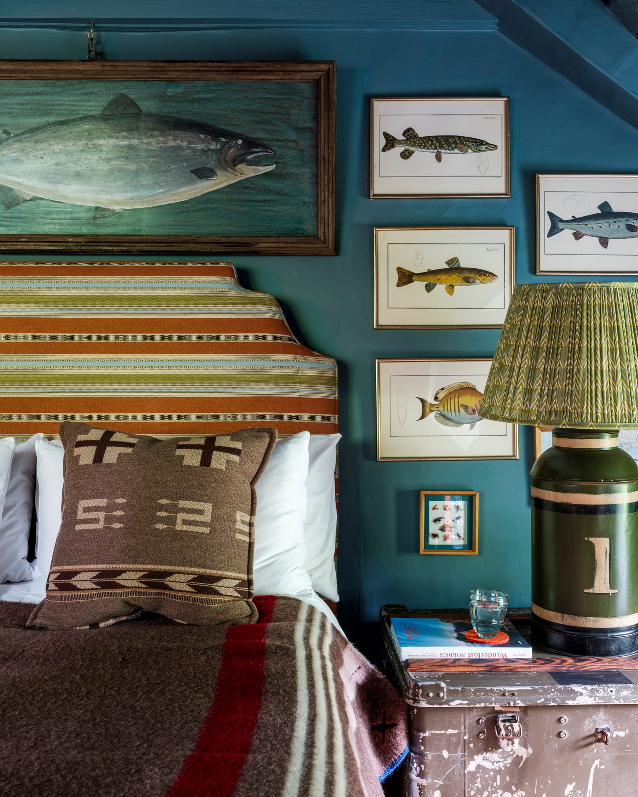 Cozy bedroom featuring deep teal walls, a striped upholstered headboard, and vintage fishing artwork framed above the bed