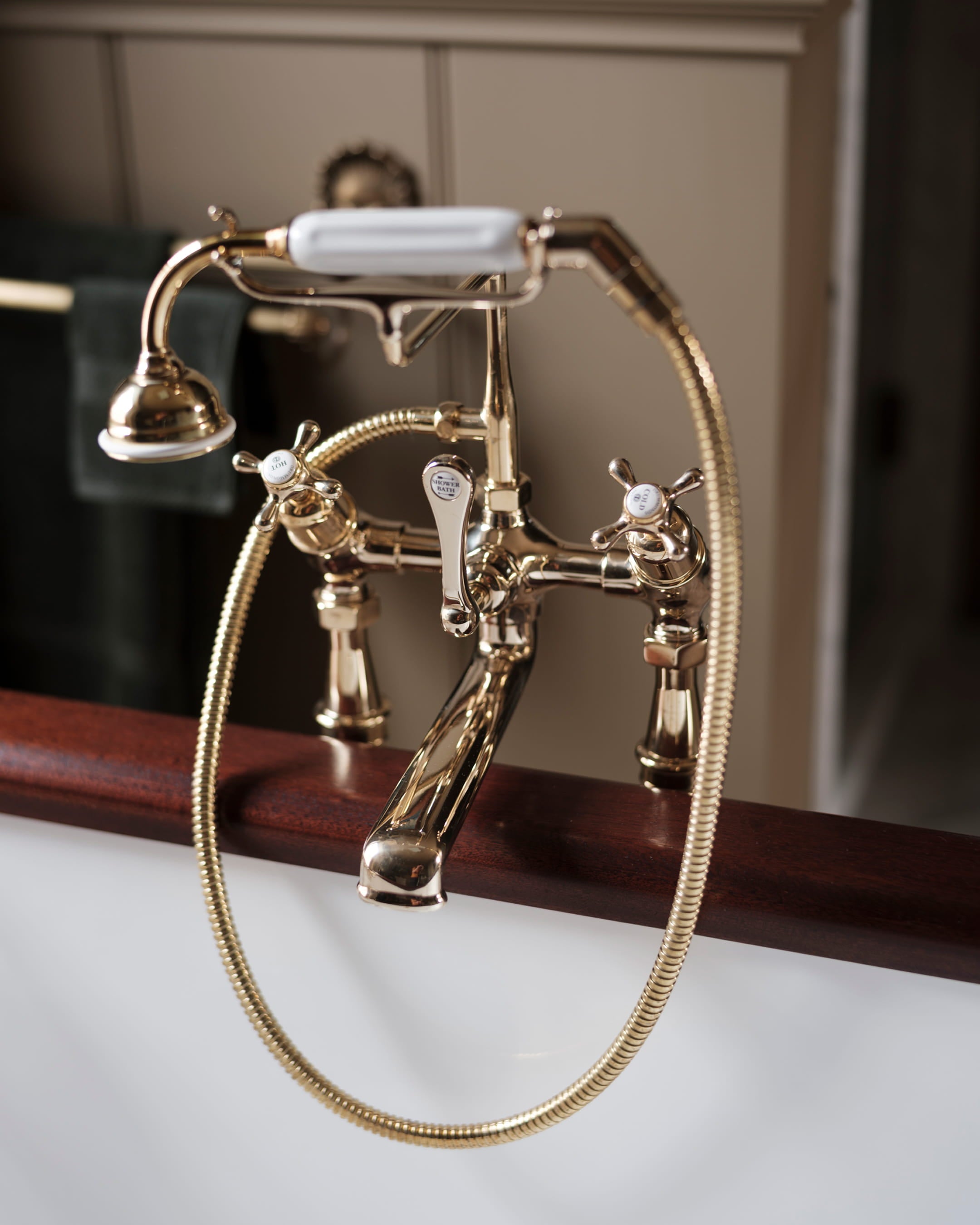 Brass bathtub faucet with white porcelain handles, set against a rich mahogany tub and soft green towels in the background