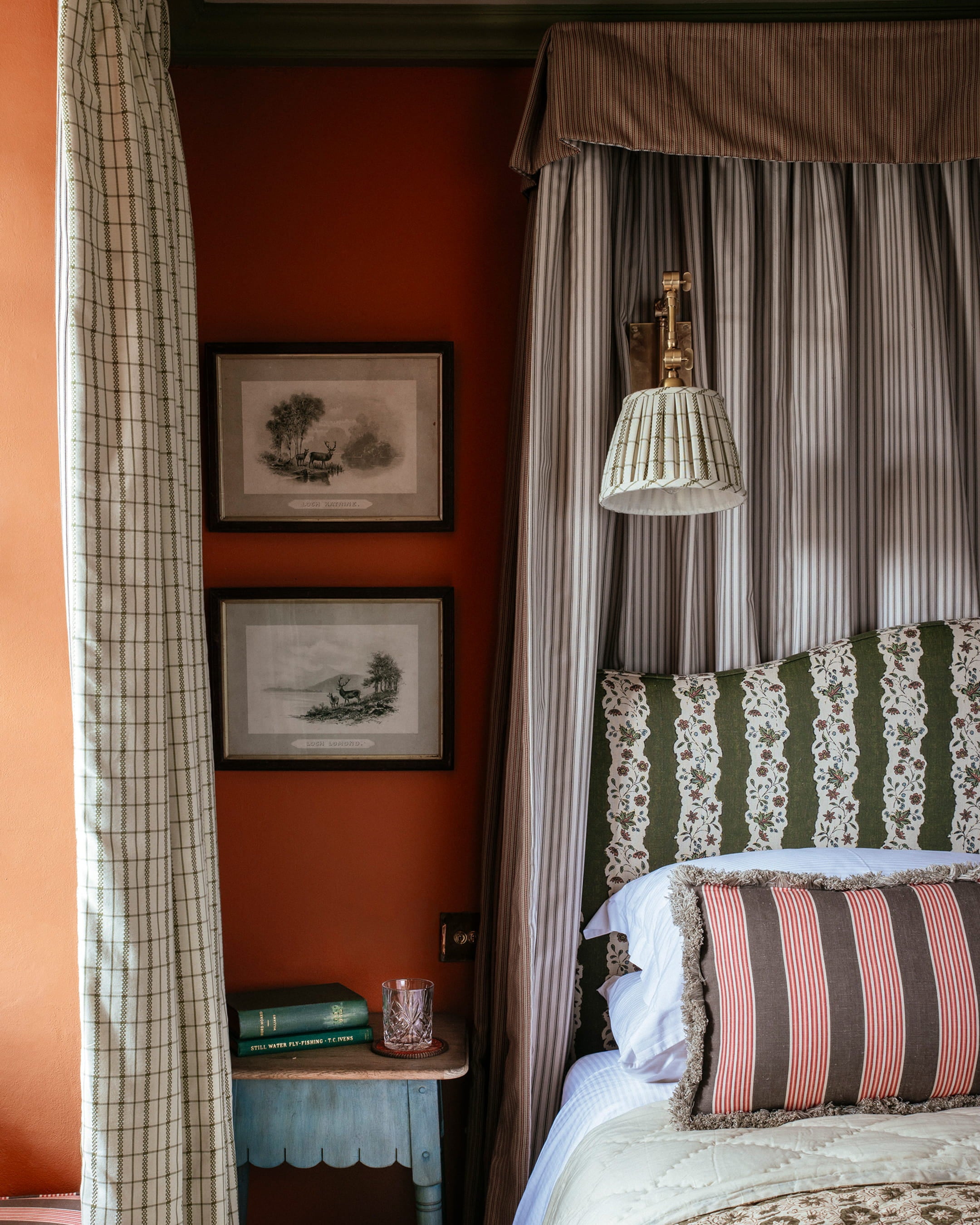 Cozy bedroom featuring burnt orange walls, a floral headboard, and a vintage bedside table with stacked books and a glass tumbler