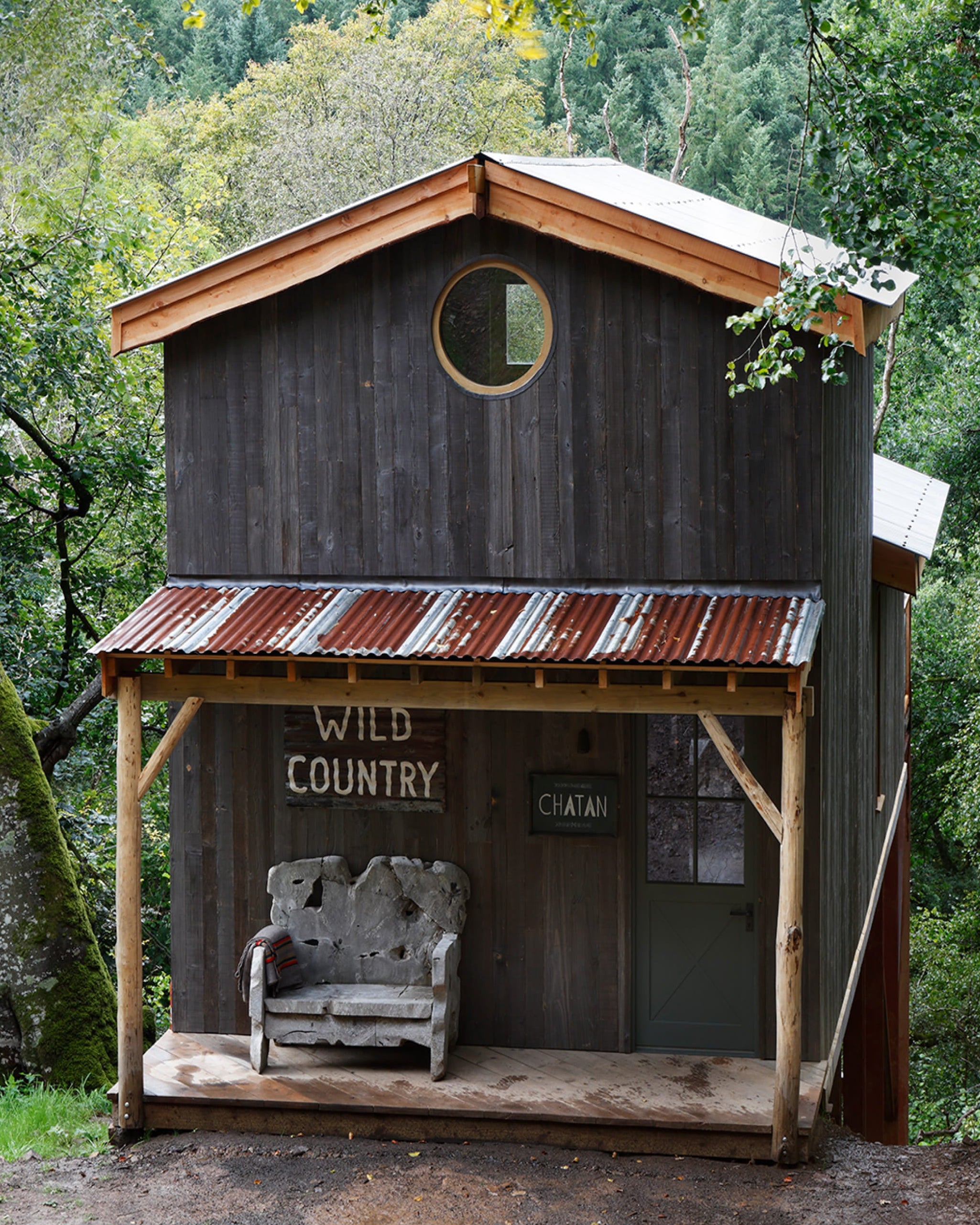Rustic cabin exterior featuring dark timber cladding, a corrugated metal roof, and a round window surrounded by greenery