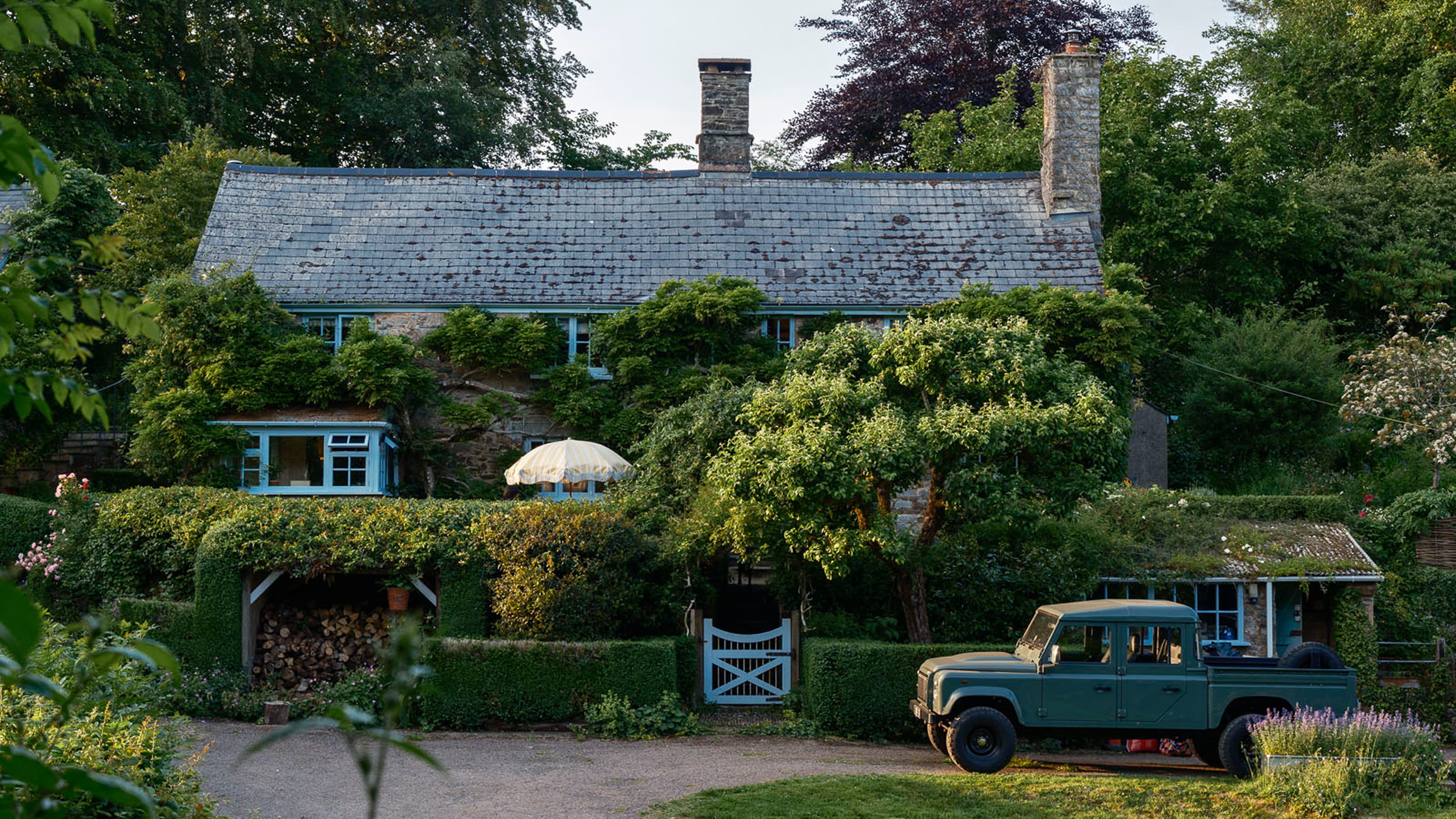 Charming stone cottage surrounded by lush greenery, featuring a slate roof, blue window frames, and a rustic wooden log store