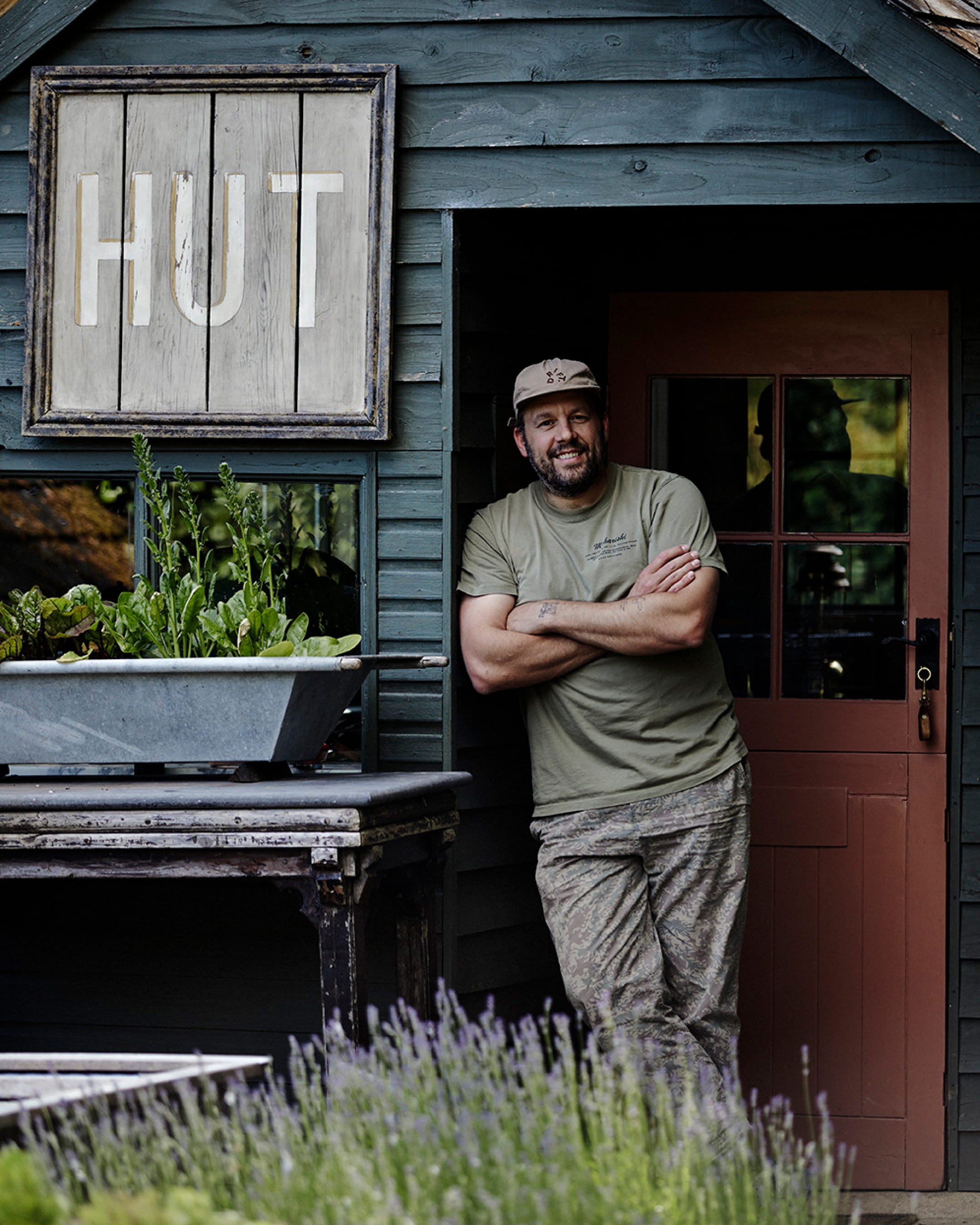 A rustic hut with dark blue wood siding, a warm terracotta door, and a large metal planter filled with fresh greens