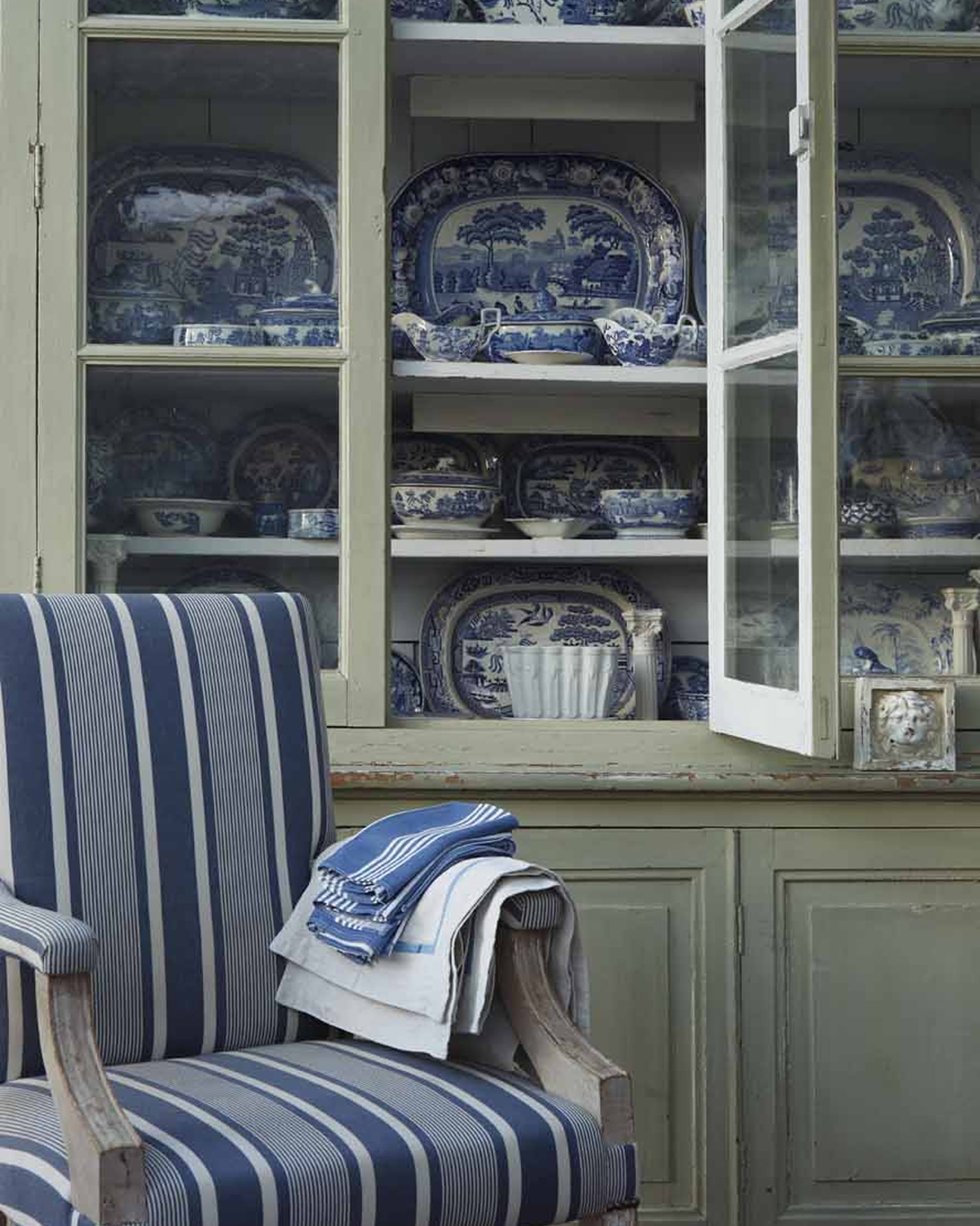 Navy and white striped armchair with a stack of linens beside a glass-fronted cabinet displaying blue and white porcelain dishes