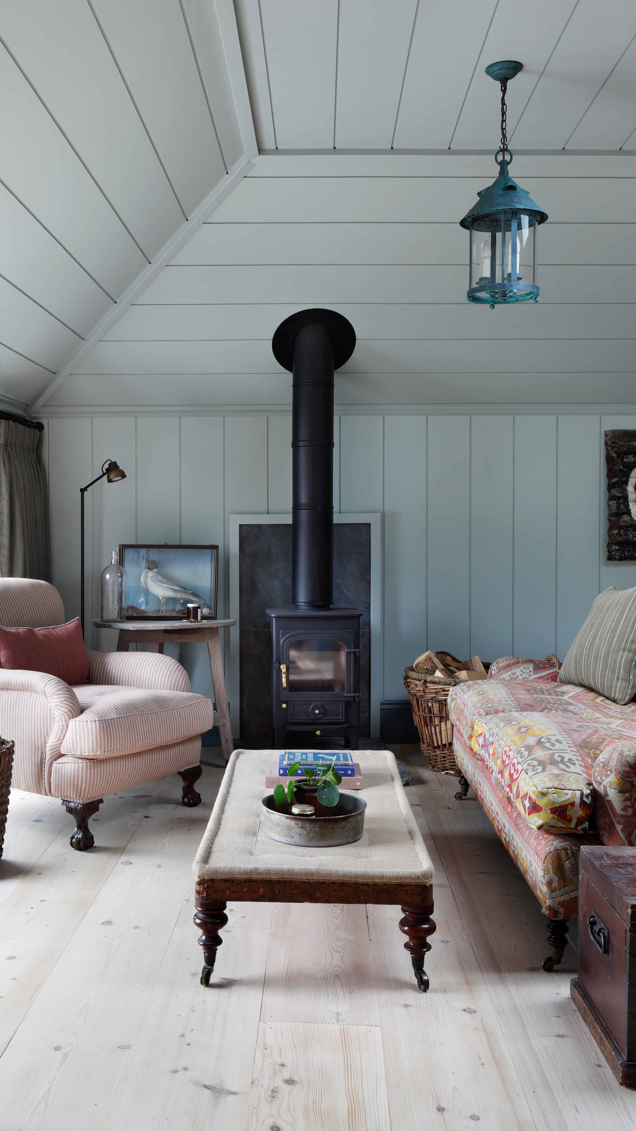 Cozy living room featuring pale blue panelled walls, a black wood-burning stove, and a vintage striped armchair with a soft cushion
