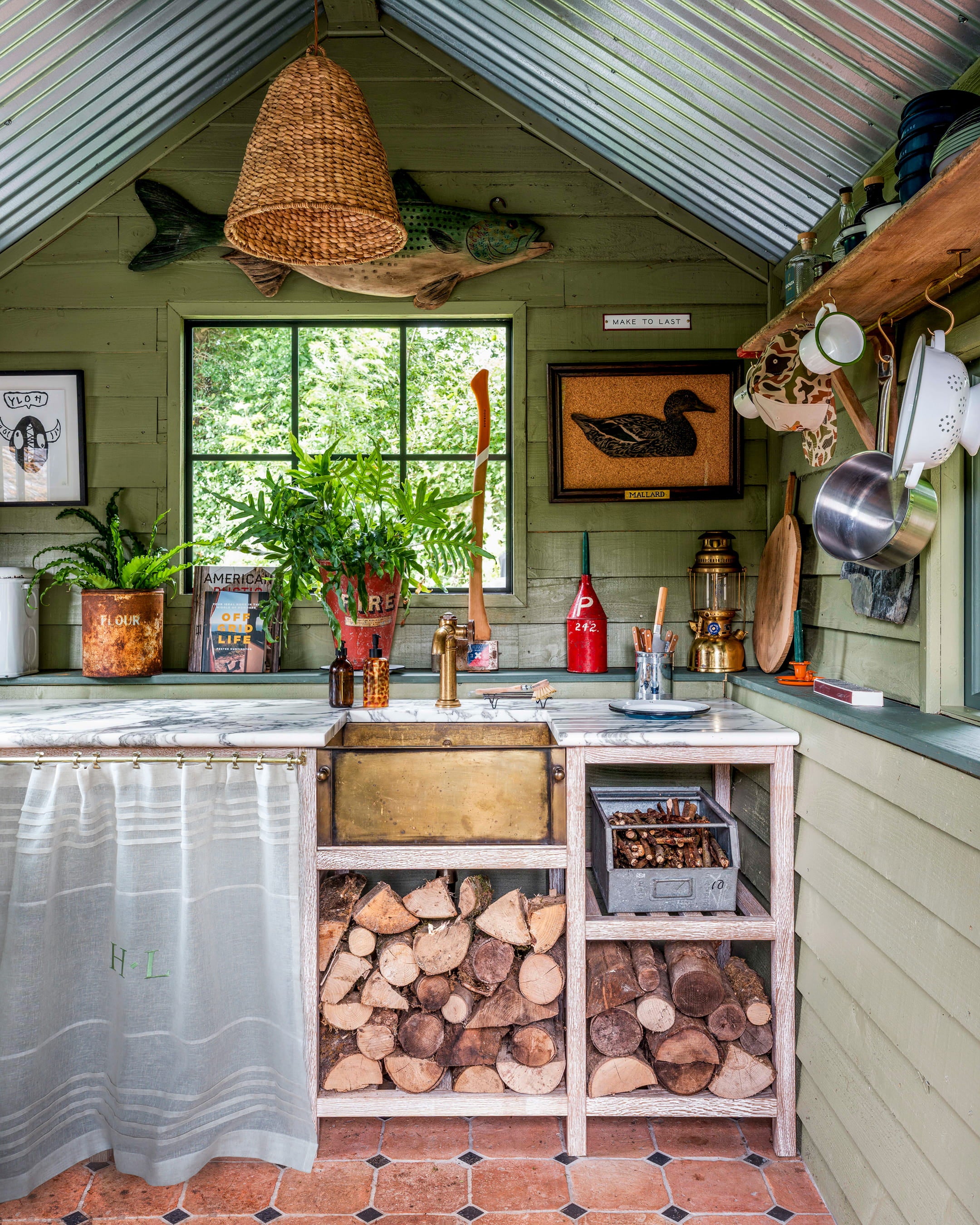 Rustic kitchen featuring sage green wooden walls, a marble countertop, and a brass sink with exposed wooden shelves and logs below