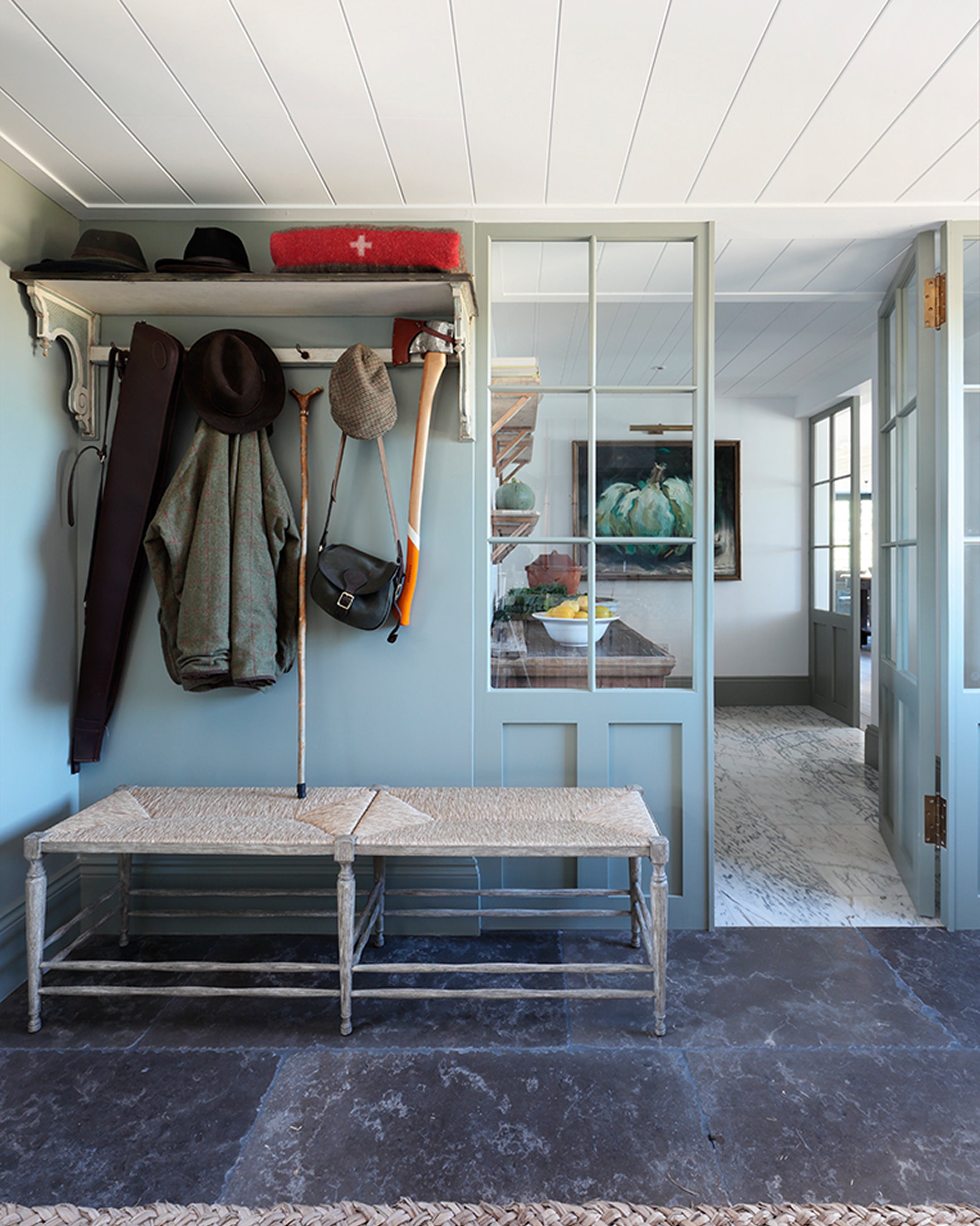 Entrance hall featuring soft blue walls, a woven bench, and slate flooring, with rustic hats and coats hanging nearby