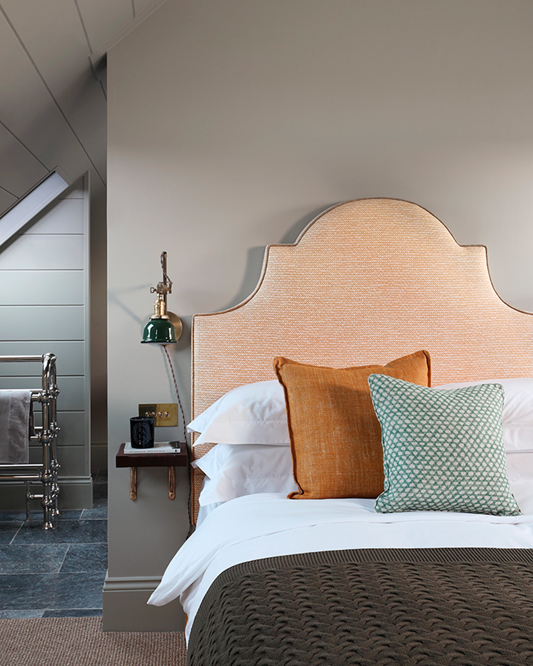 Attic bedroom featuring a woven rattan headboard, terracotta and green cushions, and slate flooring with chrome towel rail