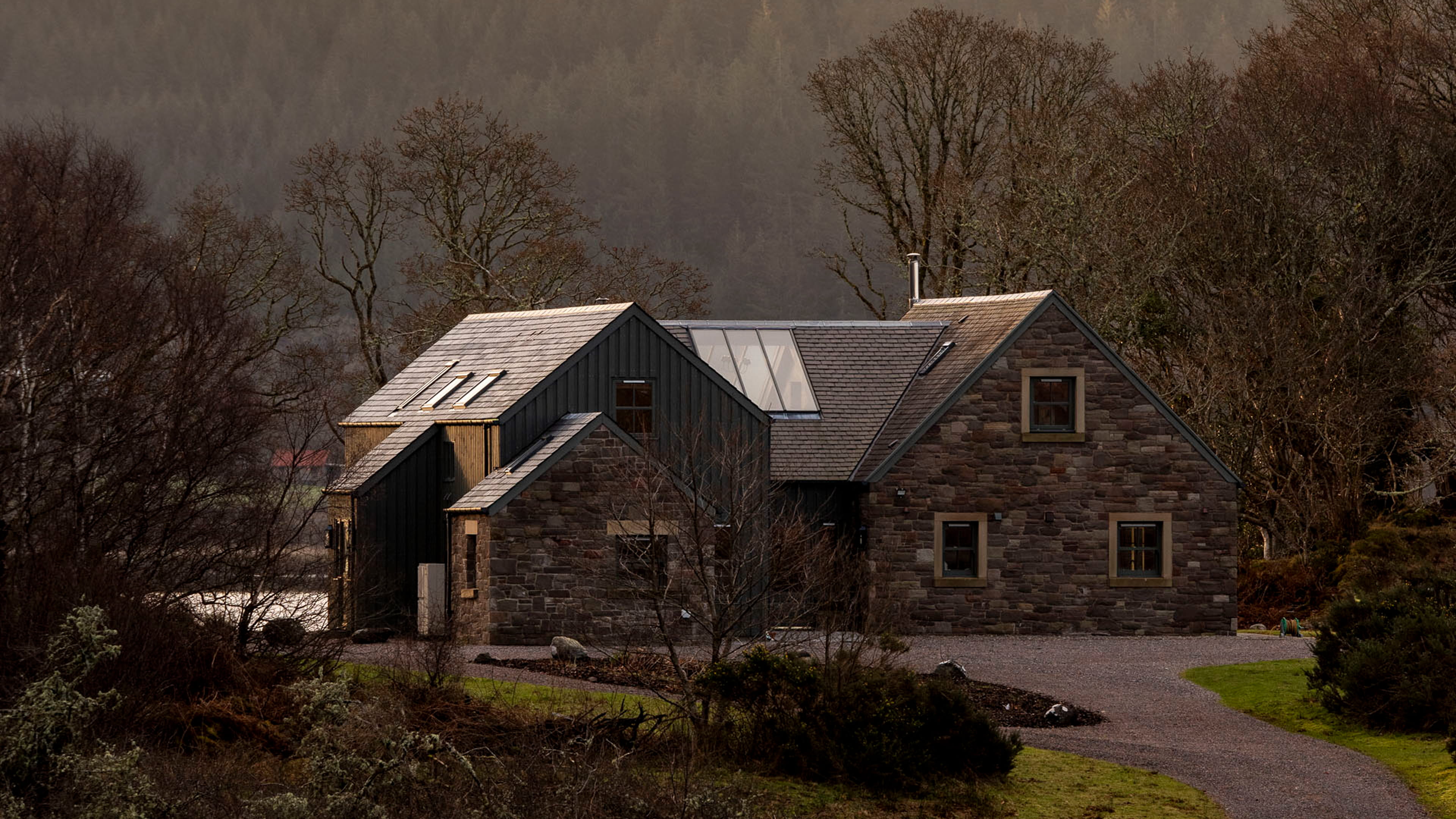 Contemporary stone-clad house with dark timber accents, large glass windows, and a sloped roof surrounded by trees