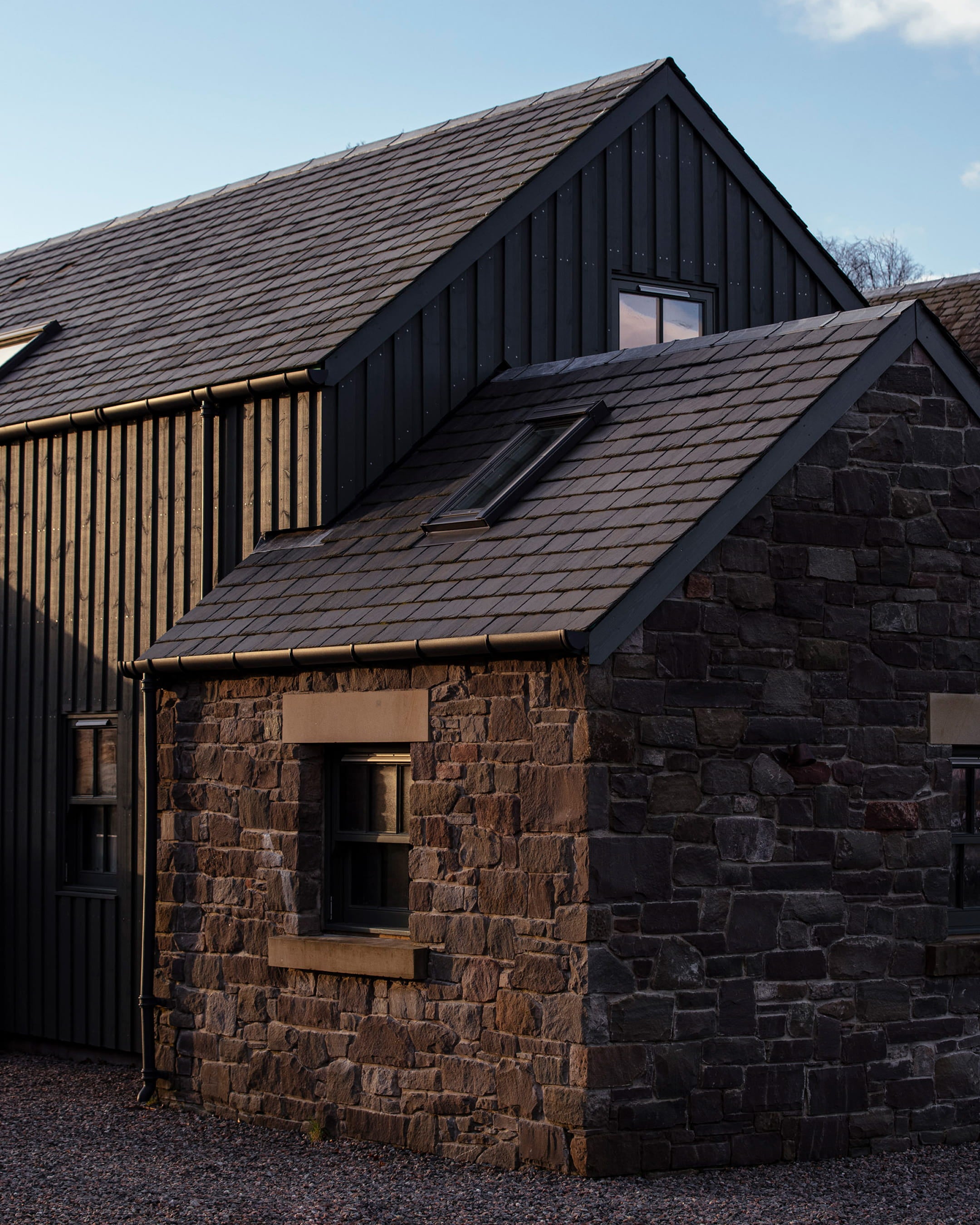 Slate grey roof and dark timber cladding on a contemporary stone cottage, featuring a large window and textured stone walls